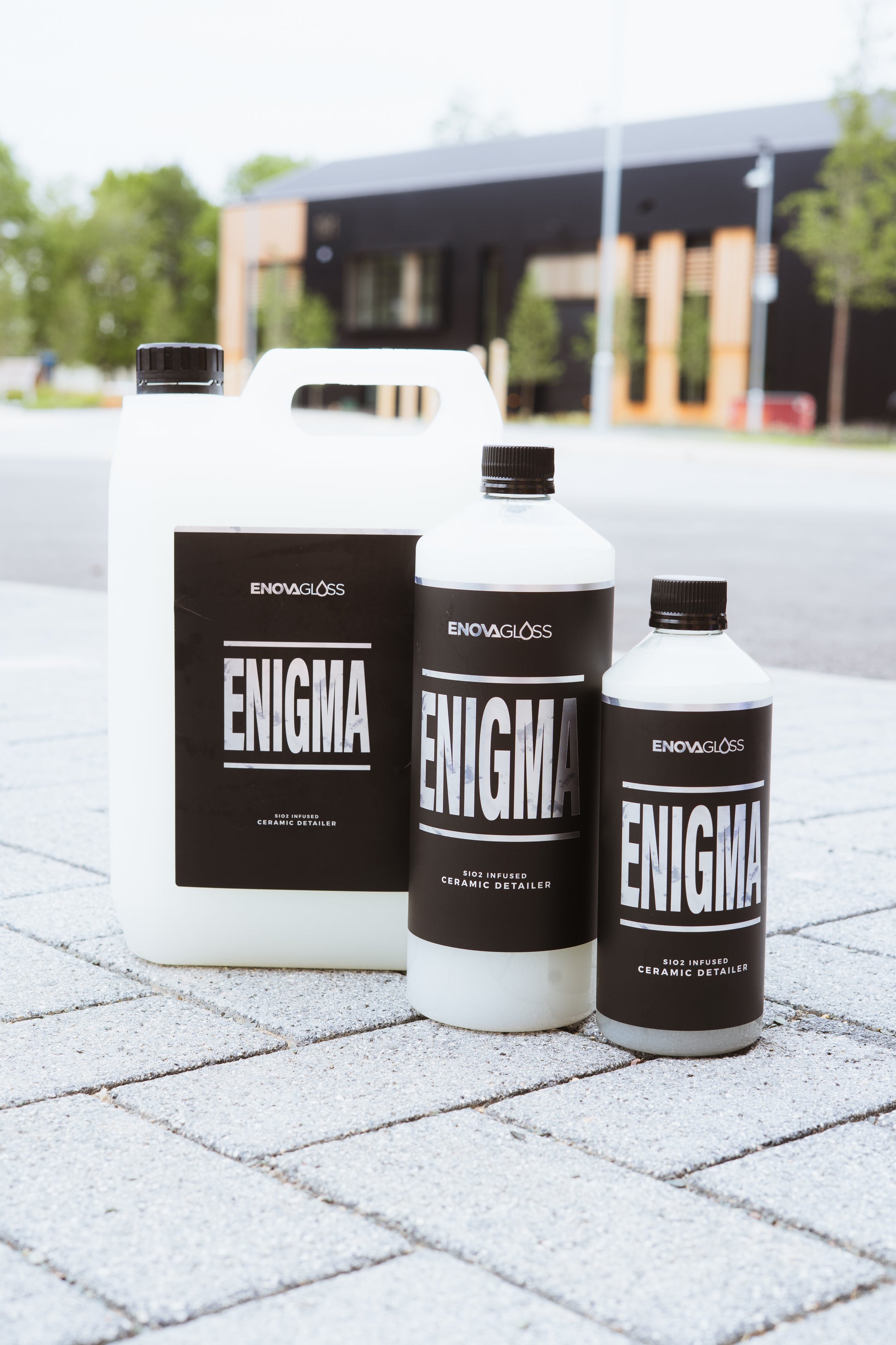 Three 'ENIGMA' bottles from the brand 'ENOVAGLOSS' on a paved surface with a blurred background.
