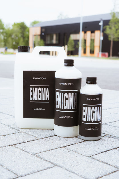 Three 'ENIGMA' bottles from the brand 'ENOVAGLOSS' on a paved surface with a blurred background.